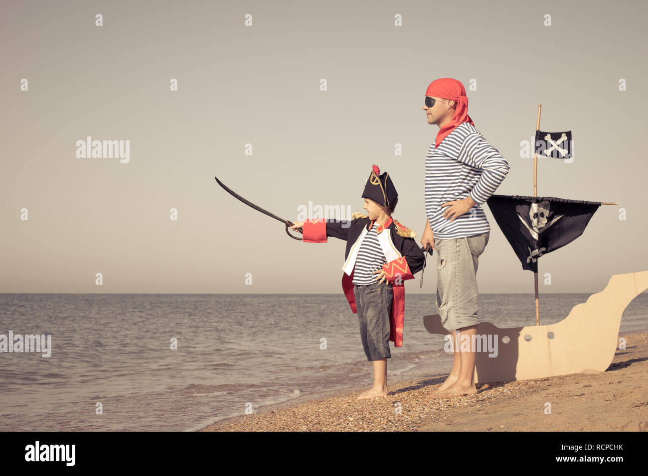Father and son playing on the beach at the day time. They are dressed in sailor's vests and pirate costumes. Concept of happy game on vacation and fri Stock Photo