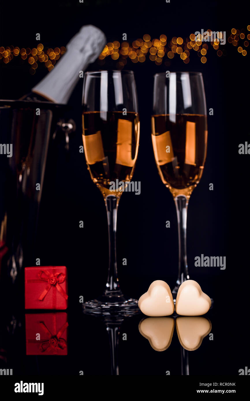 Two wine glasses, gift in a box and two heart-shaped chocolates on black background with golden lights . Love, Valentines day concept Stock Photo