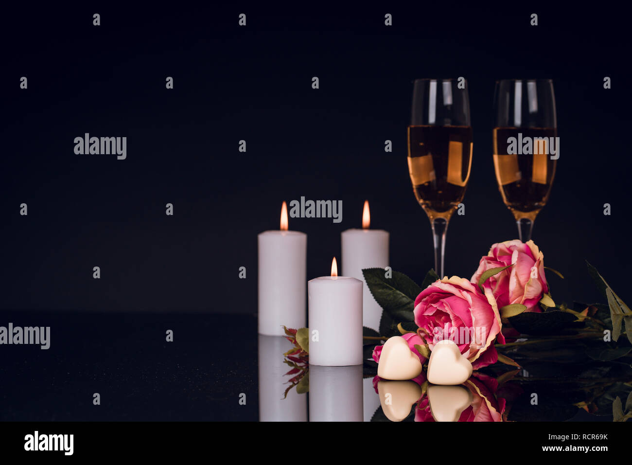 Two glasses of champagne, roses, candles and two heart shape chocolates on black background with lights in the background. Love, Valentines day concep Stock Photo