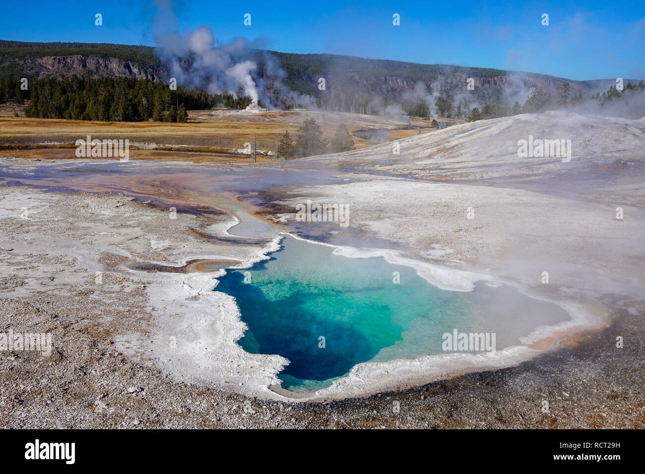 Bright blue turquoise pond (thermal feature) at Yellowstone National Park, with smoke (steam) and mountains in the background. Stock Photo