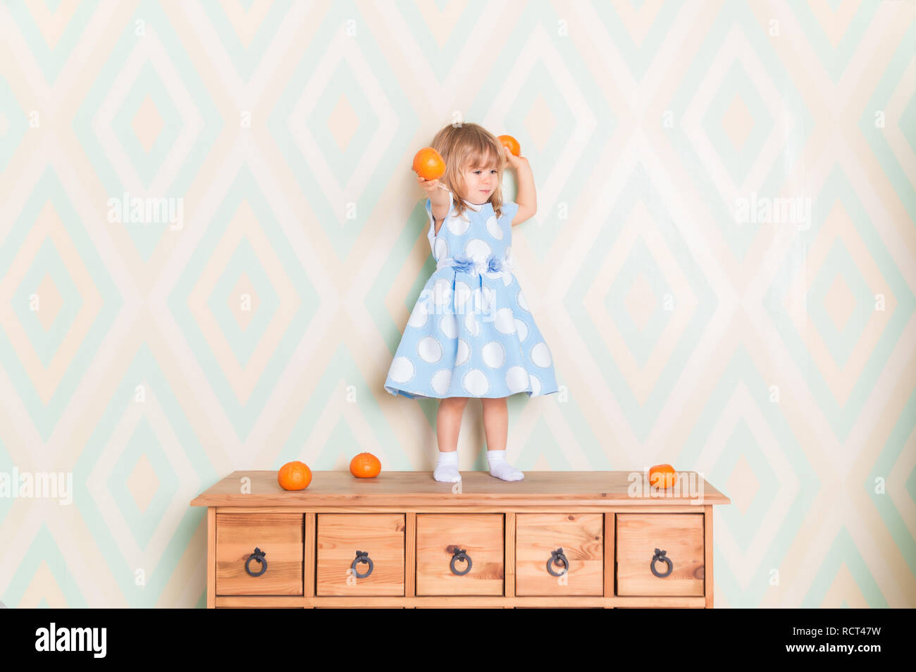 Toddler child girl in blue dress standing on wooden chest of drawers and holding fresh orange mandarins in her hands and going to throw them on rhomb wallpaper background. Winter holidays or christmas Stock Photo