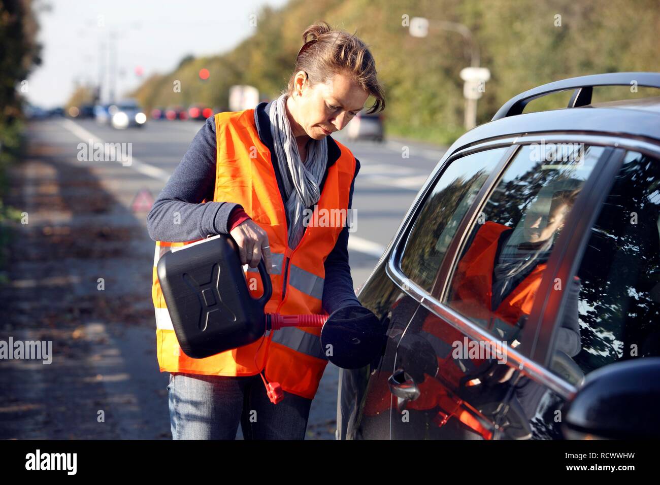 Car breakdown, female driver has stopped on the hard shoulder of a country road, wearing a reflective vest, topping up of empty Stock Photo