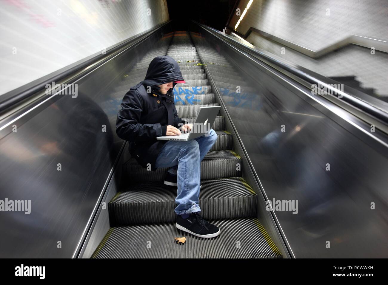 Hacker working on a laptop computer on an escalator in a subway passage at night, symbolic image for computer hacking Stock Photo