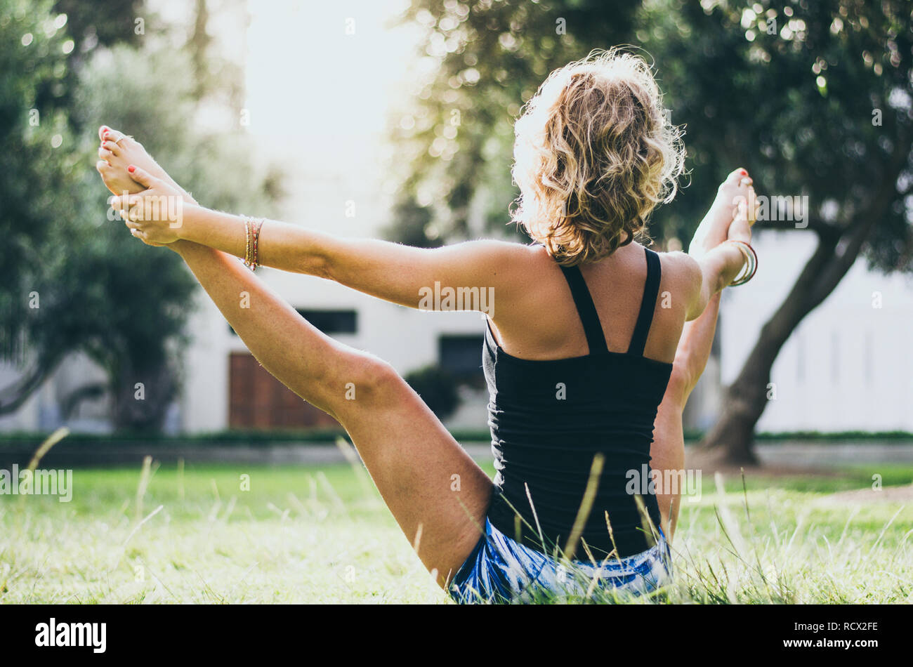Woman in jeans black tank top doing upavistha konasana yoga pose in park. Female yogi in wide angle seated forward bend holding big toes with hands in nature Stock Photo