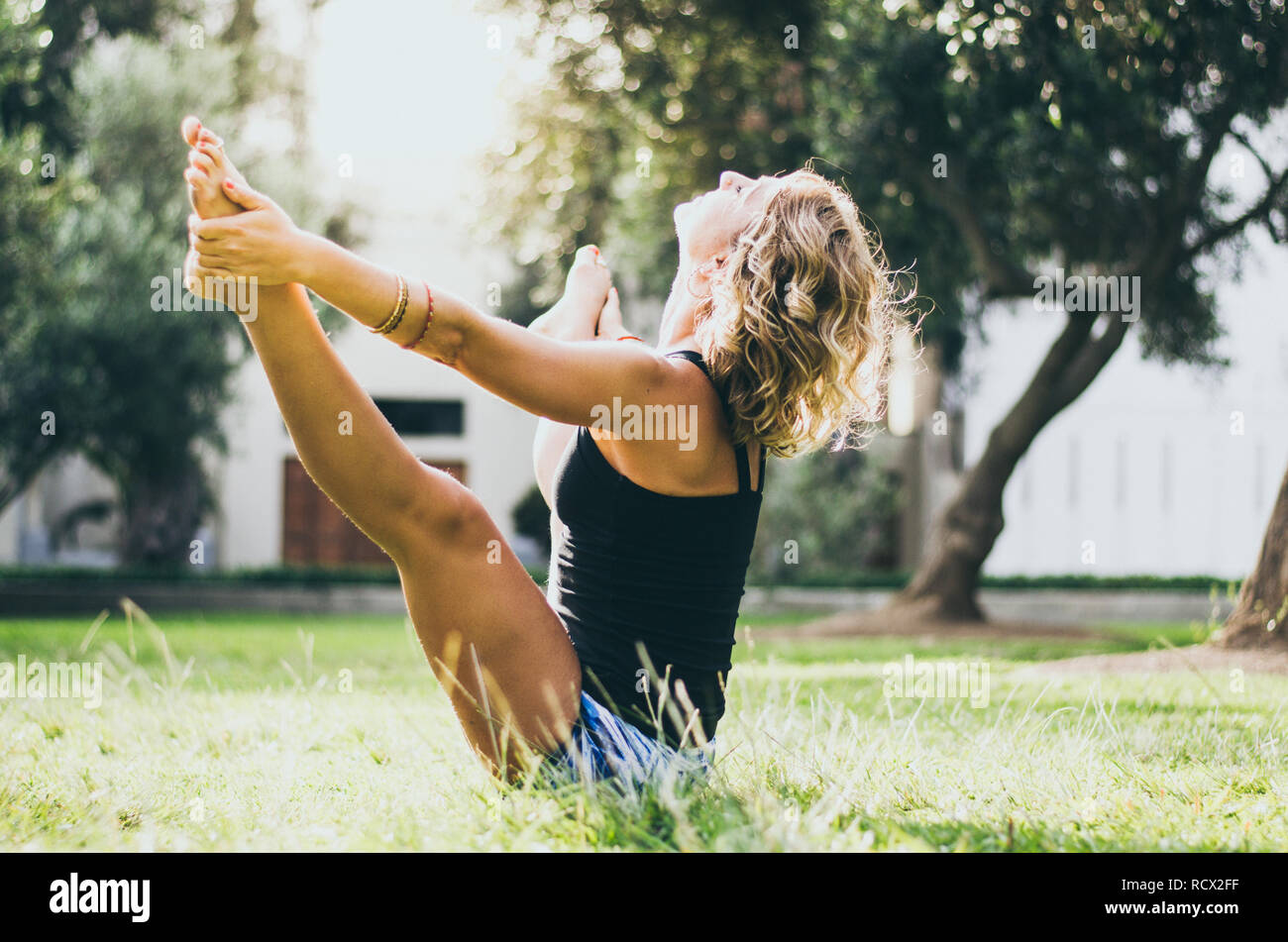 Woman in jeans black tank top doing upavistha konasana yoga pose in park. Female yogi in wide angle seated forward bend holding big toes with hands in nature Stock Photo