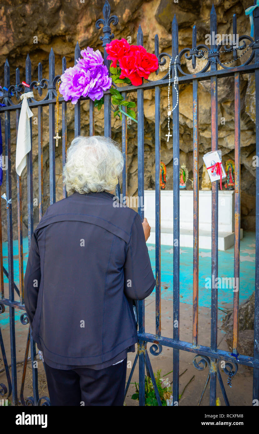 View of an elderly white haired woman viewing through an iron gate at an alter carved inside a mountain with religious icons at the historical Mission Stock Photo
