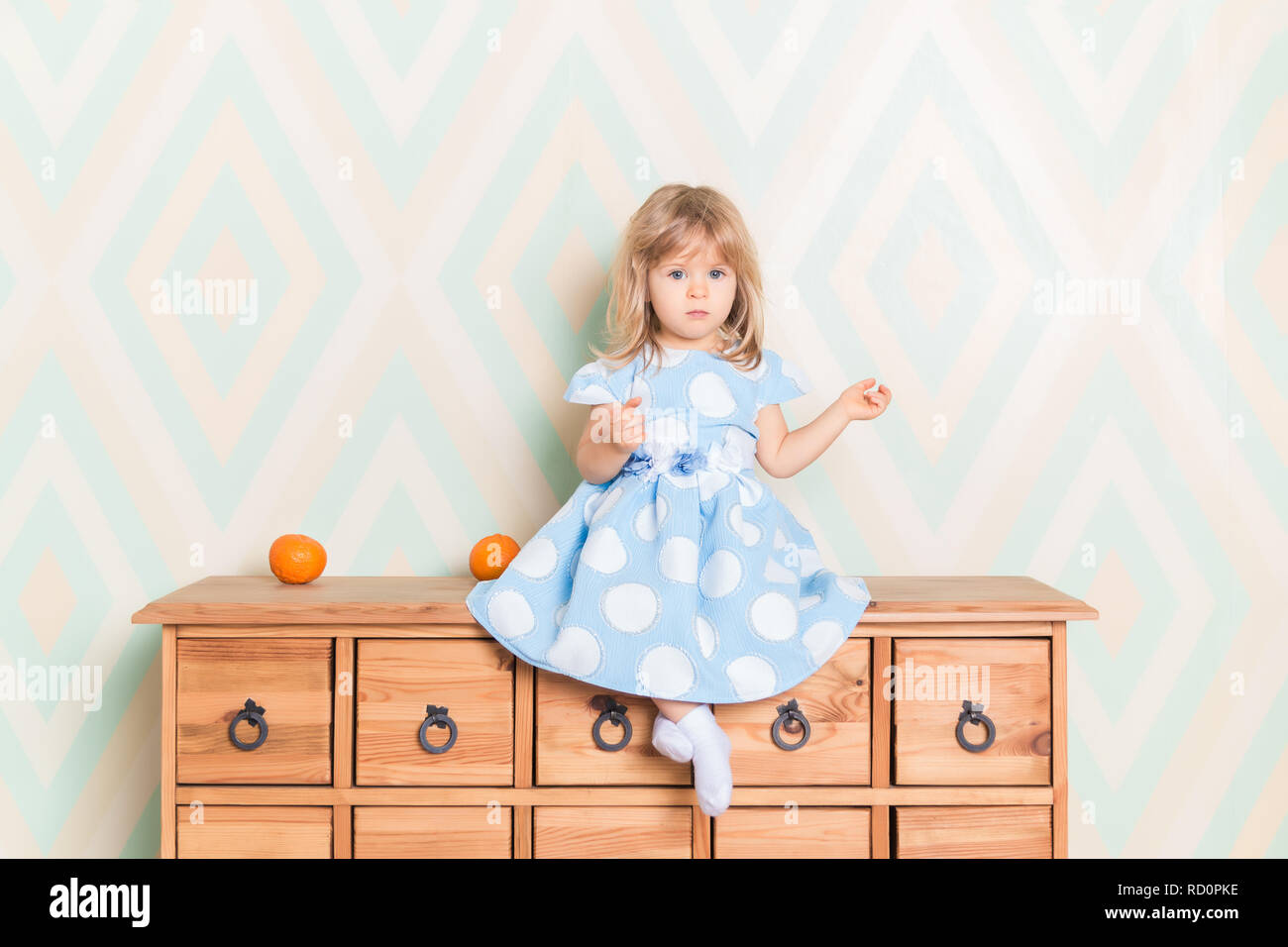 A little baby girl in her room sitting cross-legged on chest of drawers with tangerines on the rhomb wallpaper background. Child in blue polka dot dress and white socks attentively looking at camera Stock Photo