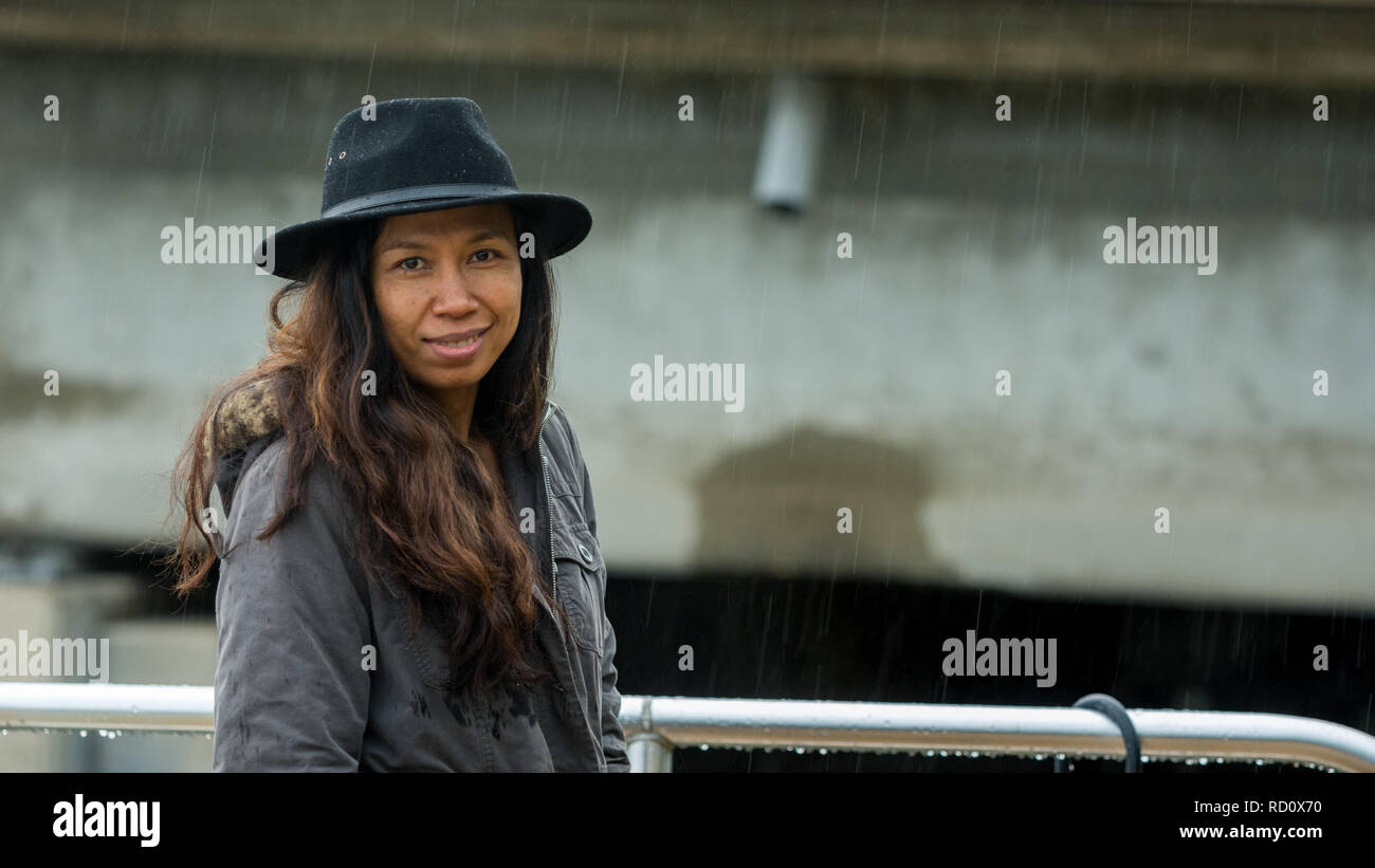 Asian woman with black hat and parka standing in the rain smiling Stock Photo