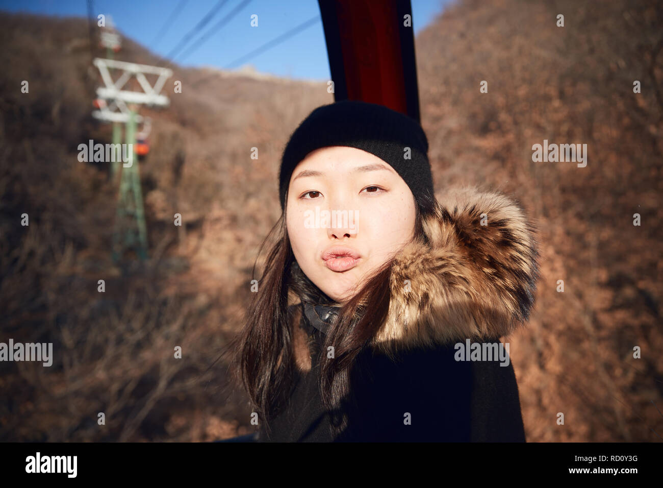 A young Chinese woman sitting inside a ski lift gondola wearing heavy winter clothing is blowing a kiss. Stock Photo