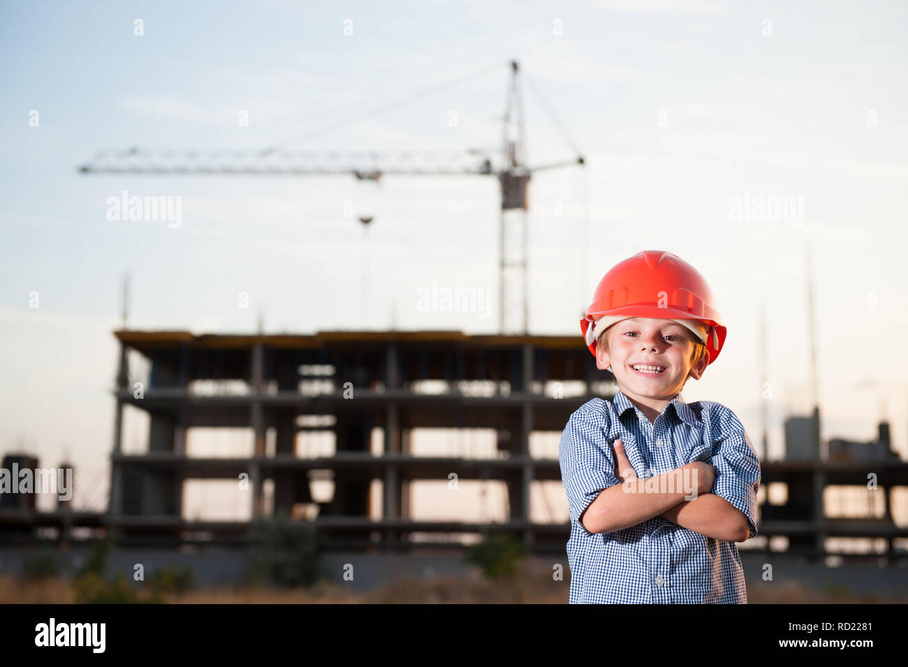 handsome happy smiling little kid in orange helmet and blue shirt standing on construction site with crane Stock Photo