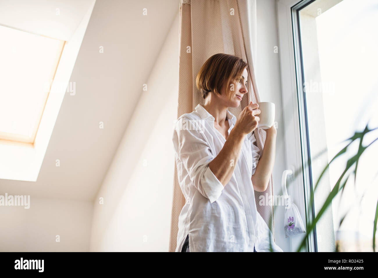 A young woman with night shirt standing by the window in the morning, holding cup of coffee. Stock Photo