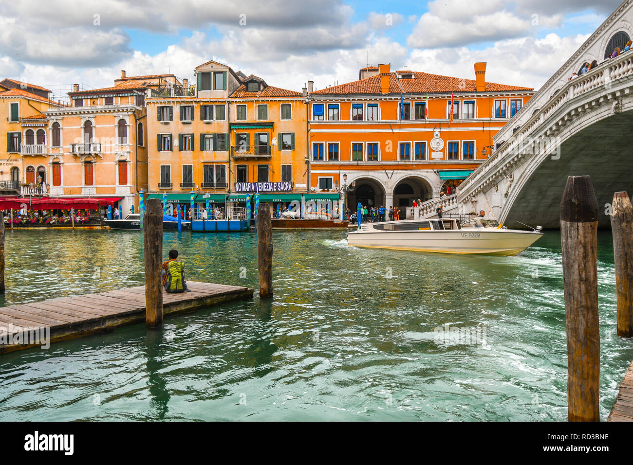 A man with a backpack sits on a dock, as tourists enjoy canal front cafes and a boat drives under the Rialto Bridge in Venice, Italy Stock Photo