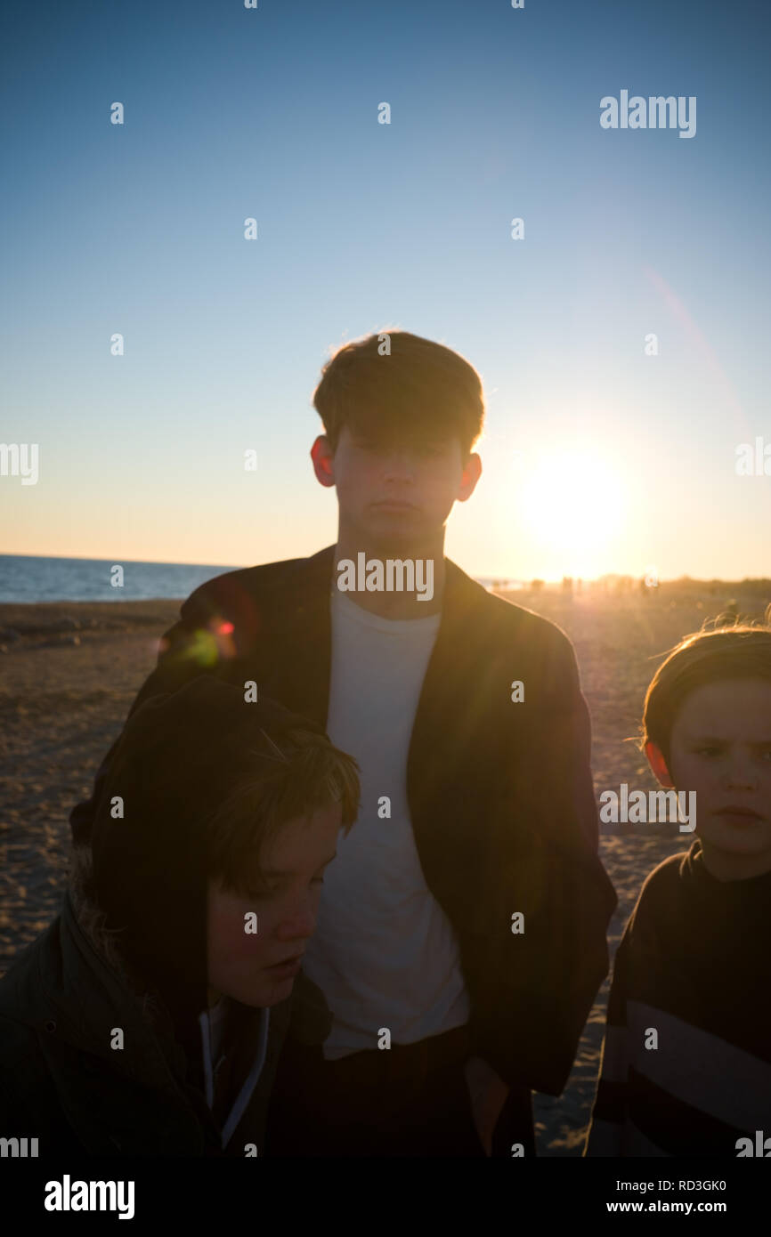 a tall dark handsome male teenager and his two younger brothers, stand with their backs to the sun on a beach at dusk in France Stock Photo