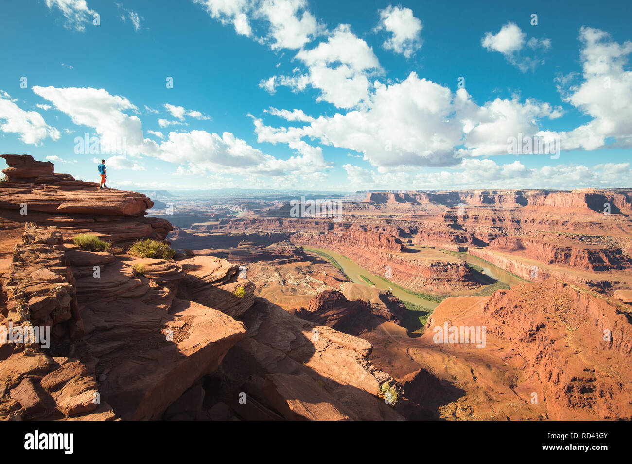 A young male hiker is standing on the edge of a cliff enjoying a dramatic overlook of the famous Colorado River, Dead Horse Point State Park. Utah, US Stock Photo
