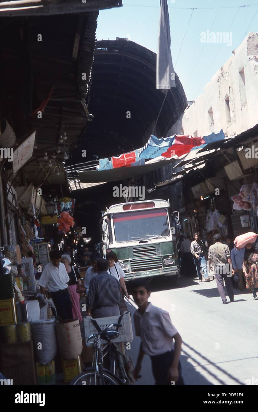 View of a busy street scene at the entrance to Al Hamidiyah souk in Damascus, Syria, June, 1994. At center a city bus travels through the market as shoppers swarm the alley's stalls. () Stock Photo