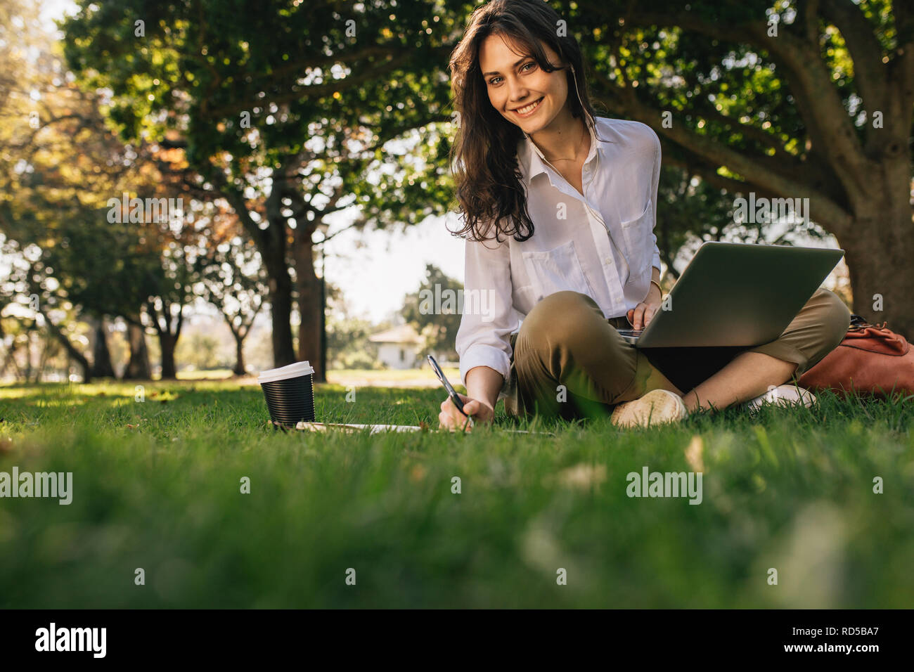 Freelancer working on laptop on green lawn in park. Beautiful woman with laptop looking at camera while working on laptop. Stock Photo