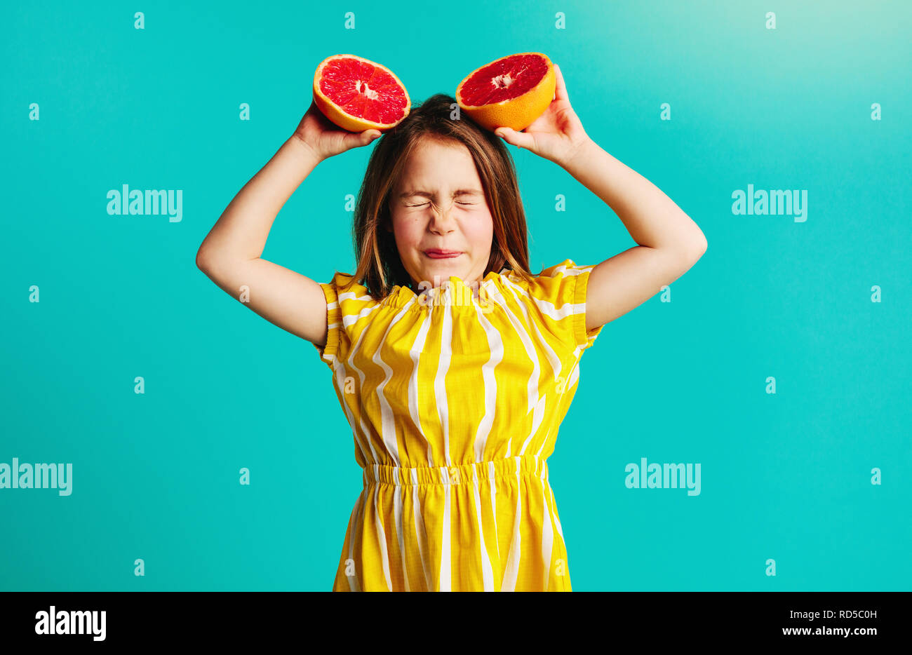 Girl holding grapefruit overhead on blue background. Cute girl does not like the taste of grapefruit. Stock Photo