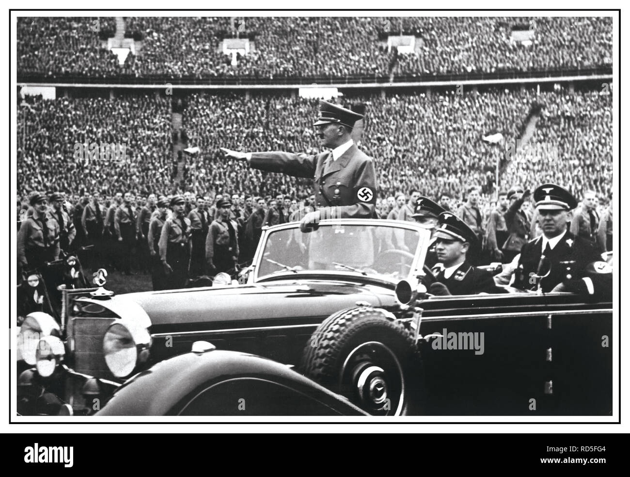 Adolf Hitler in uniform wearing swastika armband gives Heil Hitler salute to the military and crowds at a huge Nazi rally in 1938 Germany. Hitler salutes attendees at a Reichsparteitag (Reich Party Day) in Nuremberg, Germany. A uniformed Martin Bormann also in attendance sitting in rear of open top Mercedes Car Stock Photo