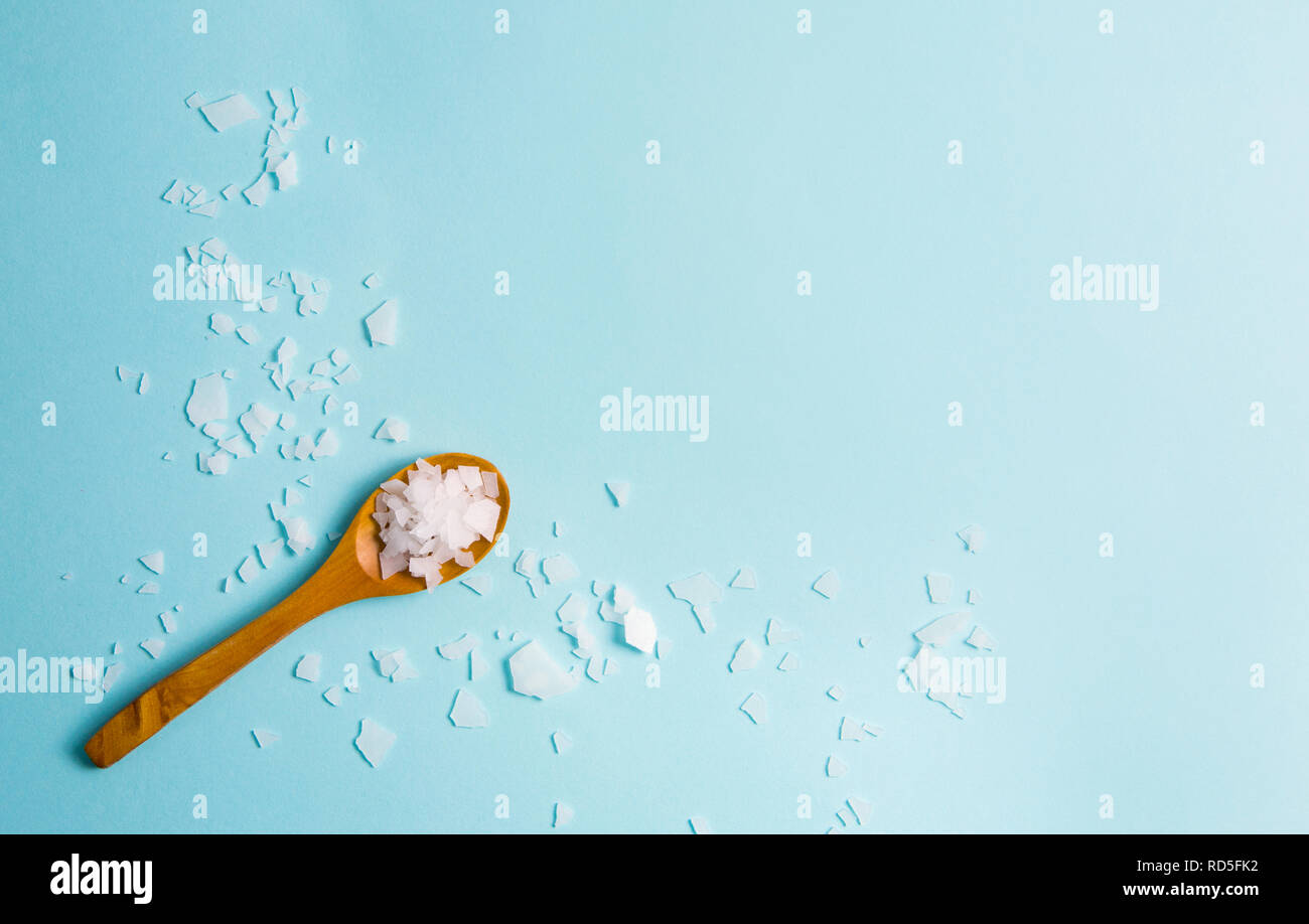 Magnesium Chloride Flakes scattered around brown wooden spoon on blue background. For making foot bath, taking a magnesium-rich bath allows full body Stock Photo