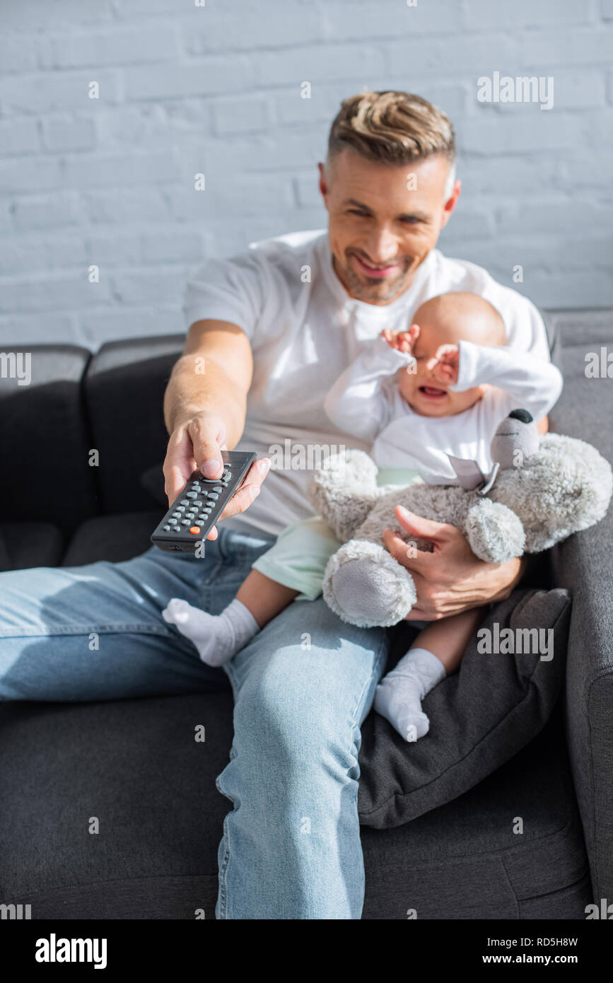 smiling father sitting on couch with baby daughter, holding remote ...