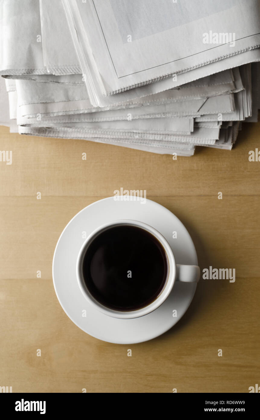 Overhead shot of a cup of black coffee with saucer on light wood laminate table with stack of newspapers. Stock Photo