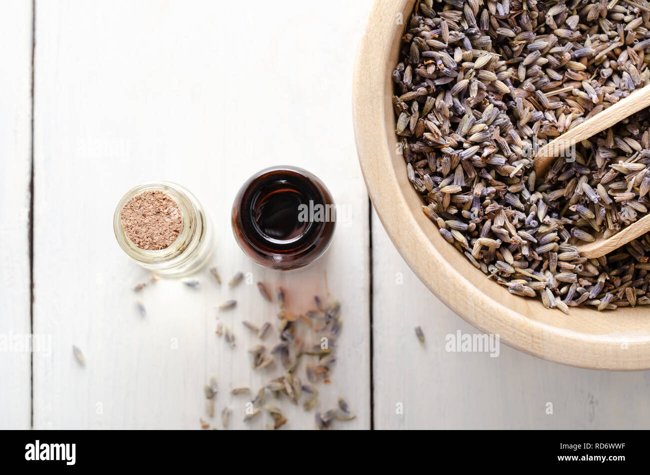 Overhead shot of a wooden bowl filled with dried lavender and two glass vials filled with oils. Further buds scattered on white wood planked table. Stock Photo