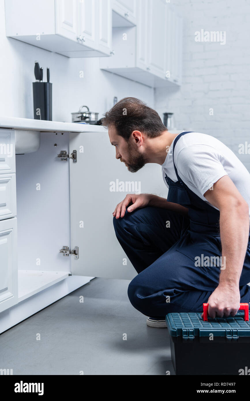 focused bearded repairman with toolbox checking sink in kitchen Stock Photo
