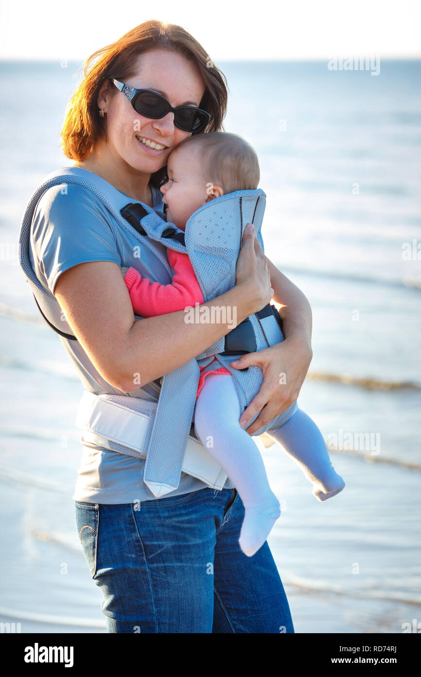Baby and mother on sea at summer day. Child in a carrier backpack Stock Photo