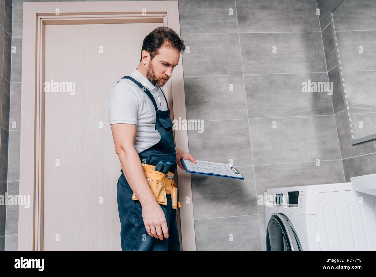repairman-with-toolbelt-and-clipboard-checking-washing-machine-in-bathroom-RD77Y8.jpg