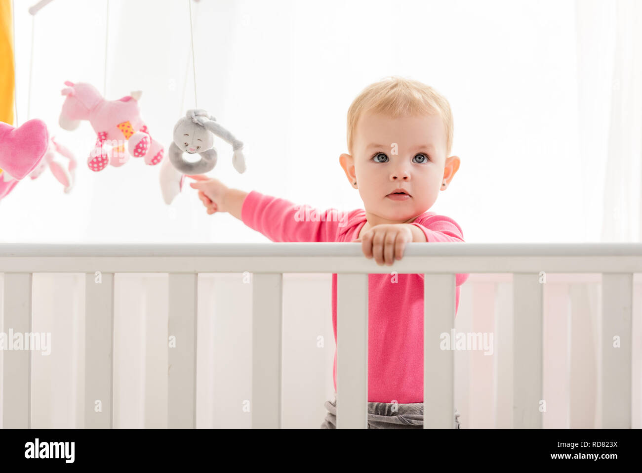Adorable Kid In Pink Shirt Standing In Crib And Touching Toys And