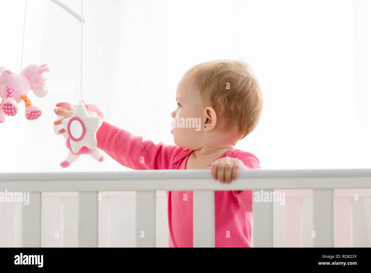 Side View Of Adorable Kid In Pink Shirt Standing In Crib And