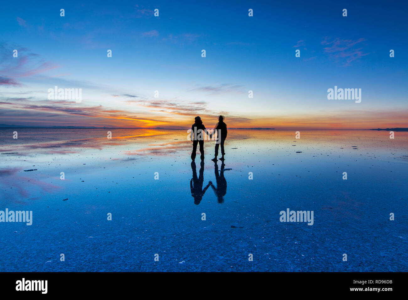 Couple looking to Sunrise at Uyuni saltflats. Sunrise over an infinite horizon in Salar de Uyuni with people reflections in water, a colorful scenery Stock Photo