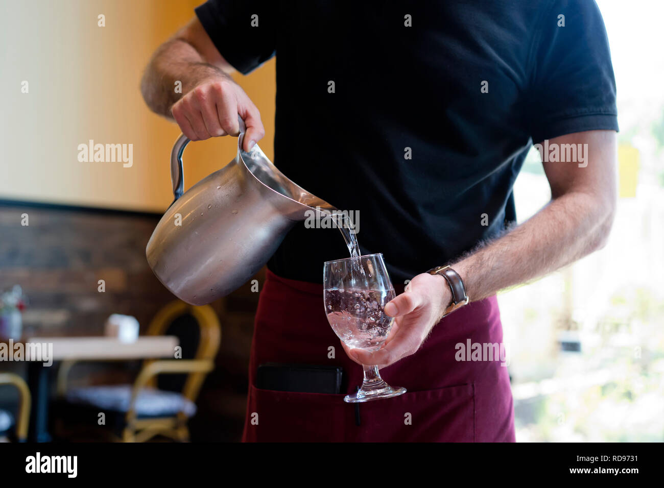 Strong slim man with muscular muscles working as a waiter in a restaurant in a black t-shirt and burgundy apron pours water from a stainless steel jug Stock Photo