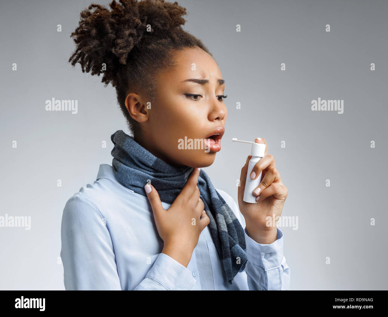 Sick woman using spray for throat. Photo of african american woman in blue shirt suffering virus of flu on gray background. Medical concept. Stock Photo