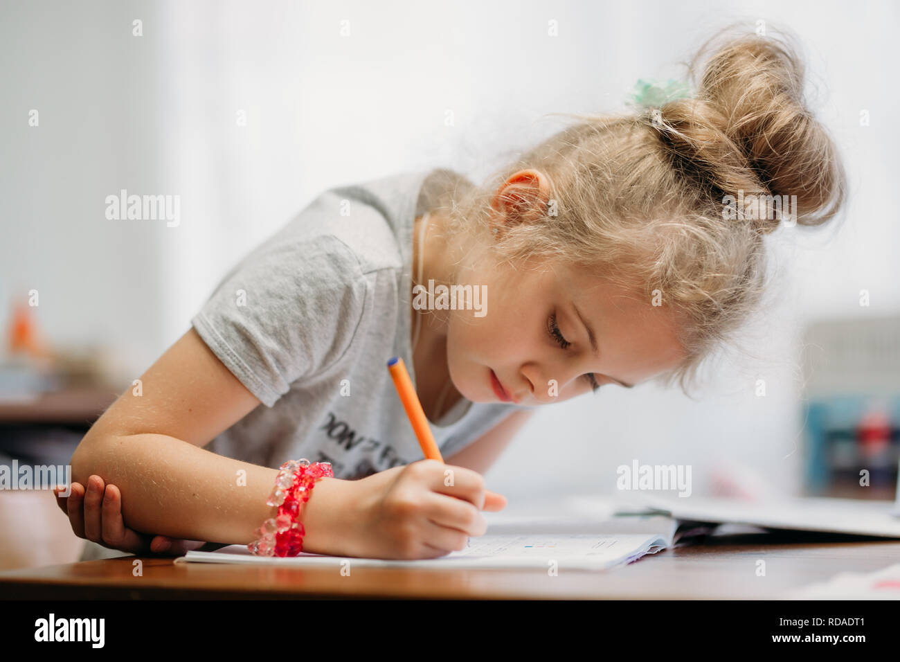 A seven-year-old girl sits at home at a table and writes in a notebook, completing a learning task or repeating lessons Stock Photo