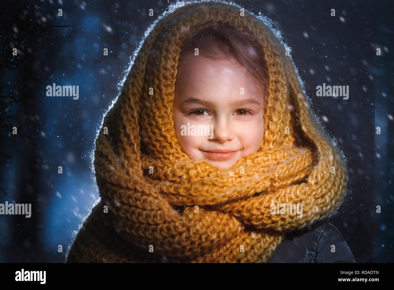 Portrait of a little charming girl in a yellow wool scarf standing outside during a snow blizzard. Stock Photo