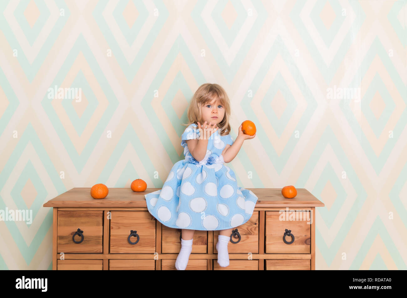 Toddler child baby girl in light blue dress sitting on the wooden chest of drawers seriously looking furrowing brows and holding fresh orange mandarins in left hand on rhomb wallpaper background Stock Photo