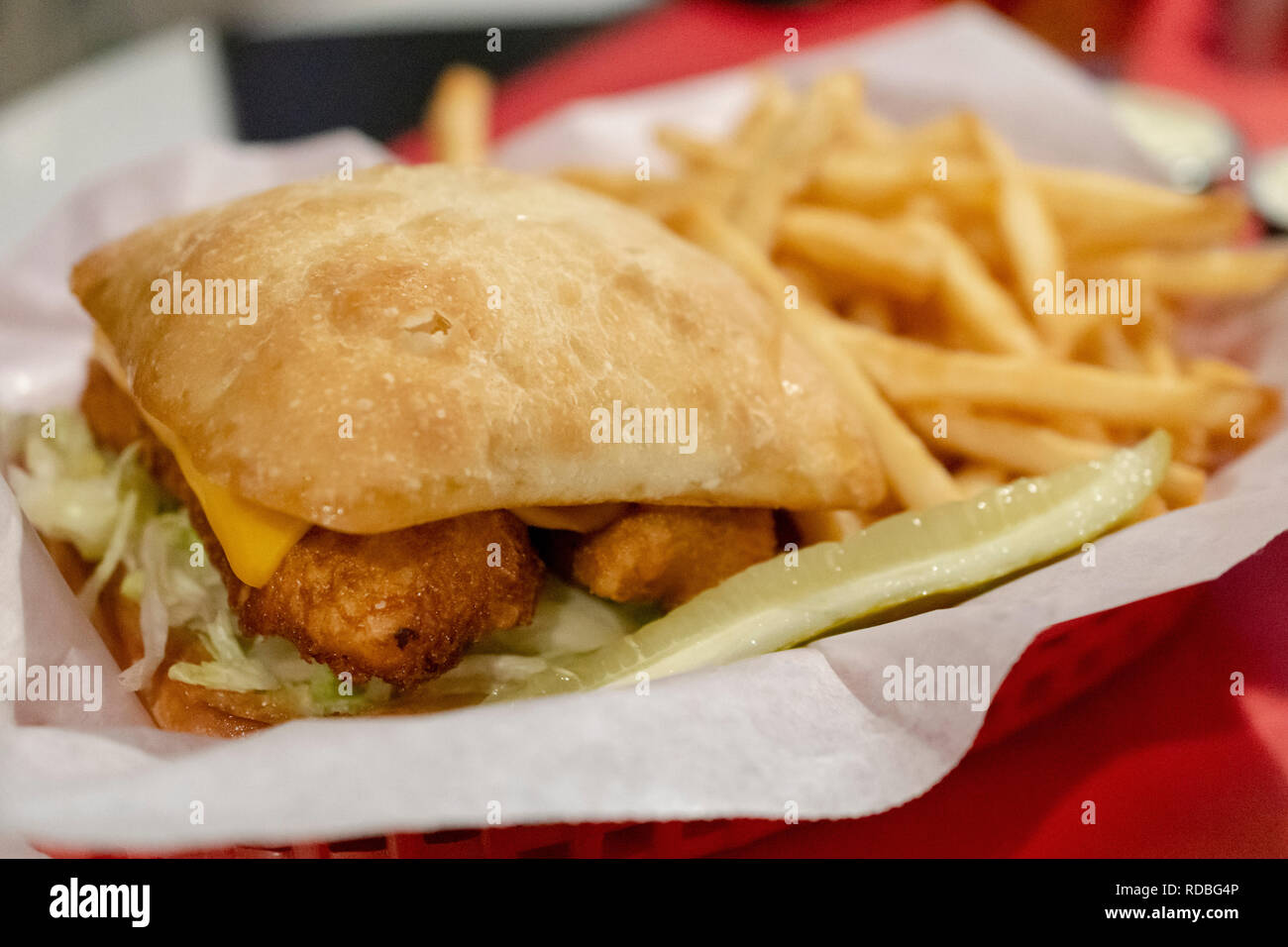 Deep fried fish sandwich on sour dough bun with cheese and lettuce.  Sandwich and fries in red basket with white paper liner.  Pickle included. Stock Photo