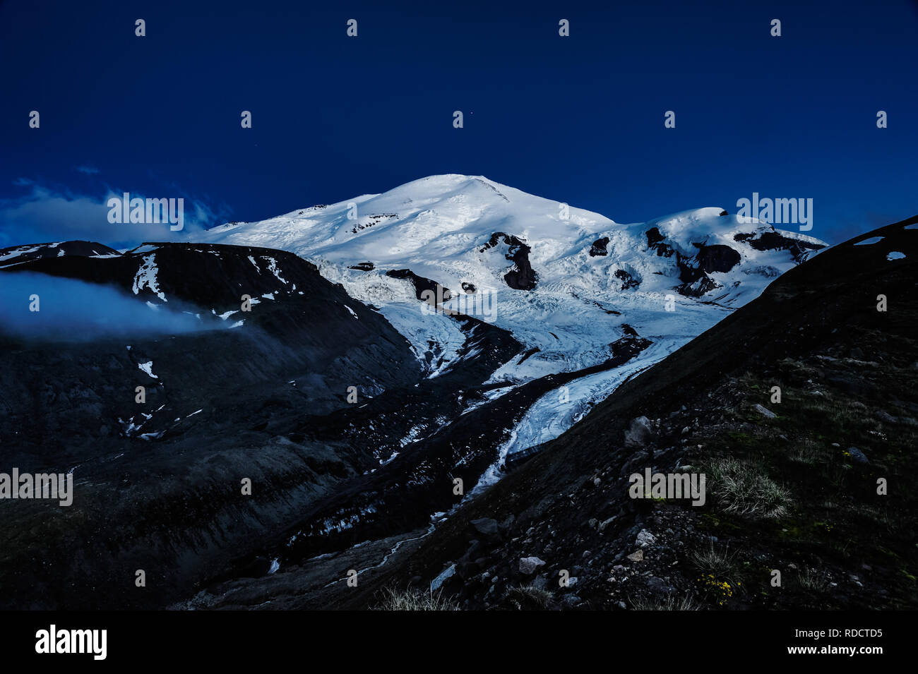 Alone snowy and icy Elbrus mountain at evening time with clear dark blue sky and one star on it. A view from a path near glacier moraine. Stock Photo