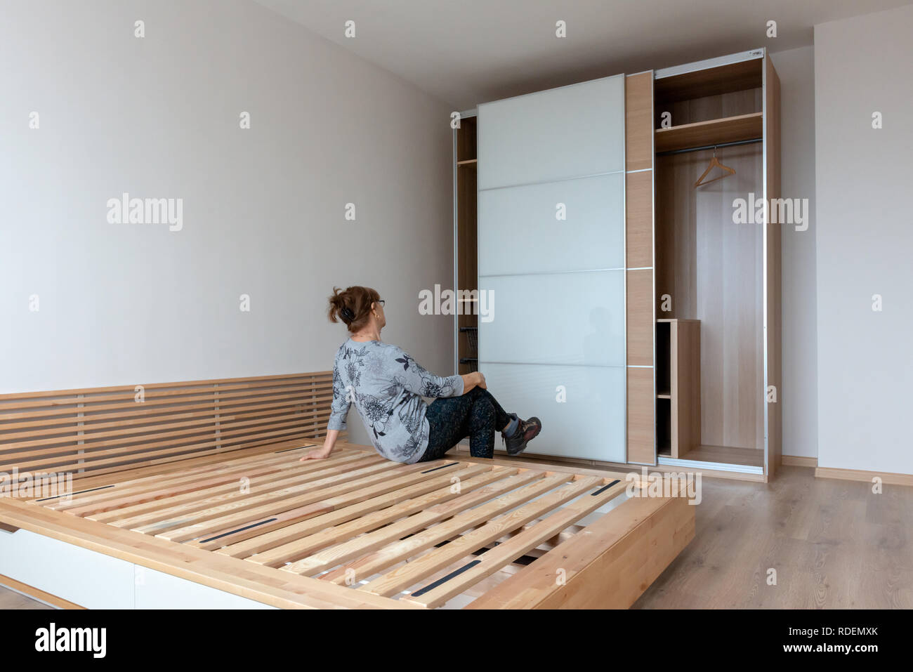 Woman admiring new wardrobe in newly furnished duotone bedroom while seating on wooden bed. New home concept. Stock Photo