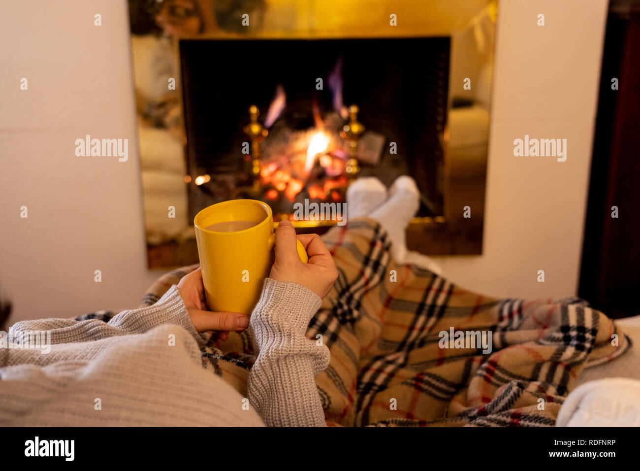 Close up image of woman sitting under the blanket by cozy fireplace warming up her feet in woolen socks relaxing with cup of hot chocolate drink at ho Stock Photo