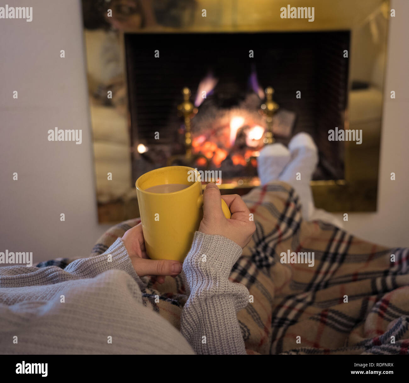 Close up image of woman sitting under the blanket by cozy fireplace warming up her feet in woolen socks relaxing with cup of hot chocolate drink at ho Stock Photo
