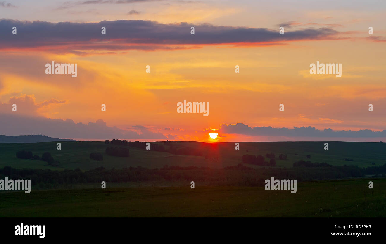 The sun sets on the horizon, the sky and the clouds are illuminated by the orange rays of the sun, summertime, the slopes of the hill are covered with. Stock Photo