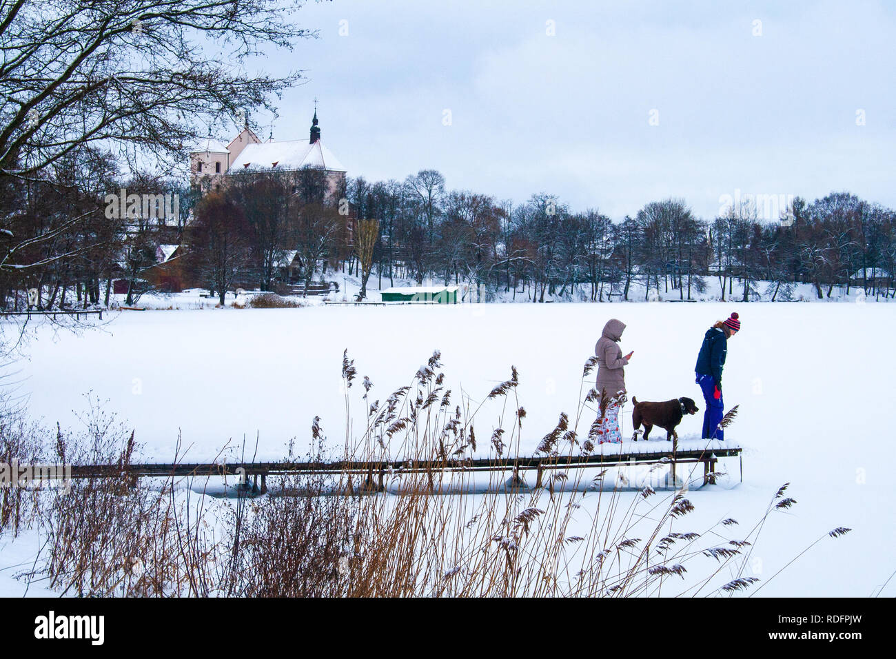 People with dog on a pier with snow on the frozen lake, forest and church in the background Stock Photo