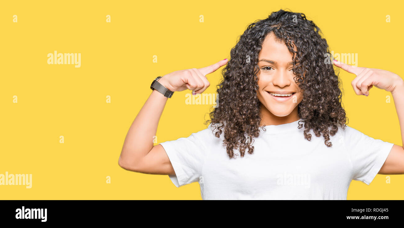 Young beautiful woman with curly hair wearing white t-shirt Smiling pointing to head with both hands finger, great idea or thought, good memory Stock Photo