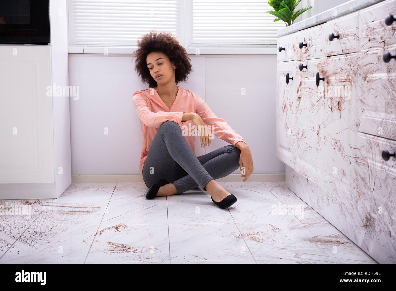 Portrait Of A Sad Young Woman Sitting On Floor With Spilled Food On The Kitchen Stock Photo