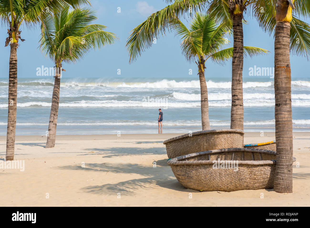 My Khe beach with a figure of a young traveler, traditional Vietnamese coracles (round wicker boats) lying on the sand, palm trees and coming waves. Stock Photo