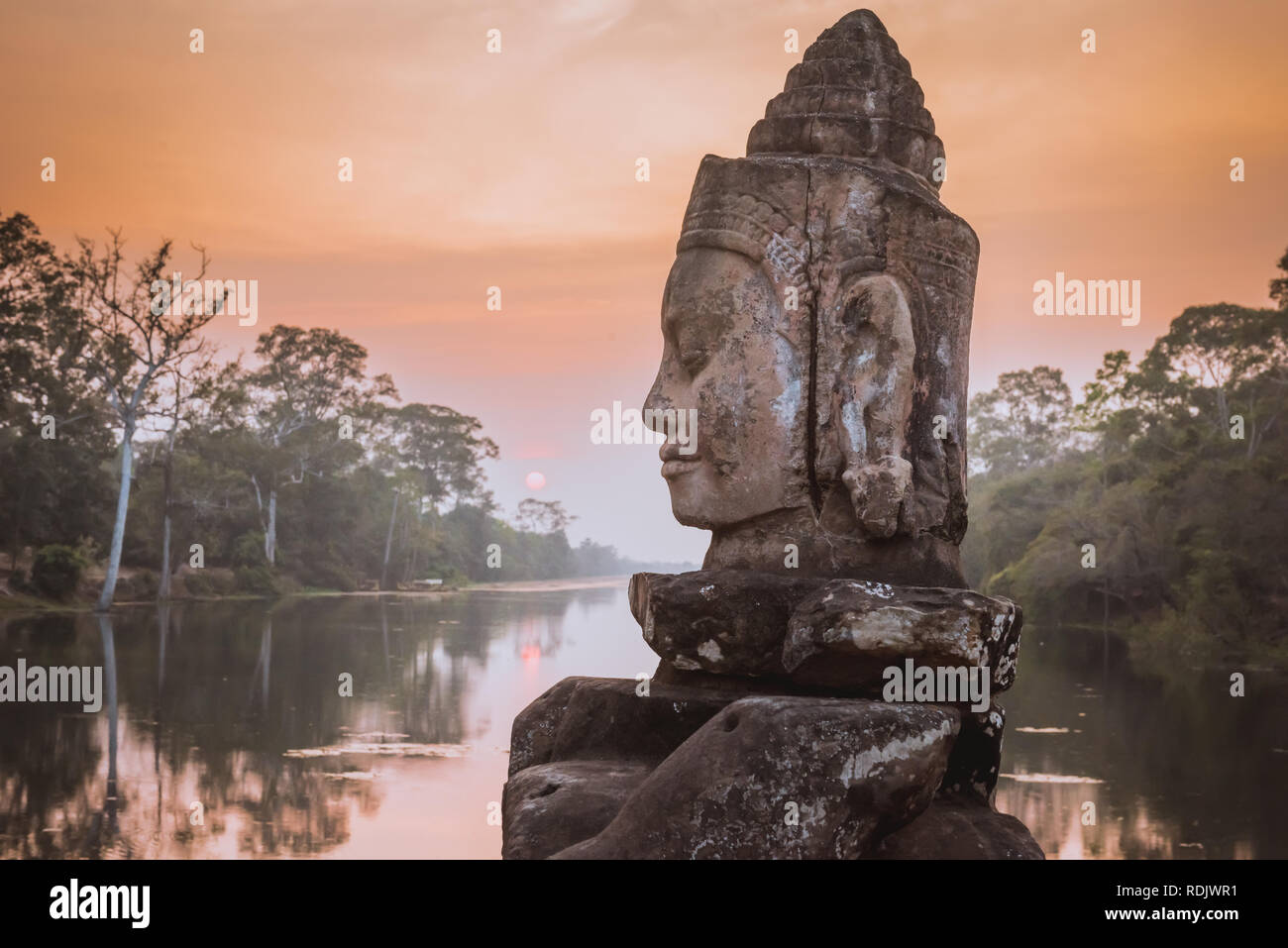 Stone Asura on causeway near South Gate of Angkor Thom in Siem Reap, Cambodia. Beautiful sunset over ancient moat in background. Angkor Thom is a popu Stock Photo