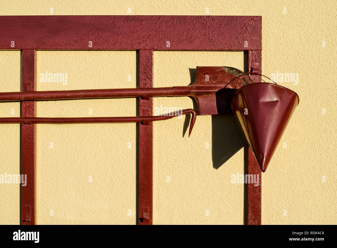 Red firefighter shield with spade hook and a cone of sand on a bright Sunny day on a background of yellow rough walls Stock Photo