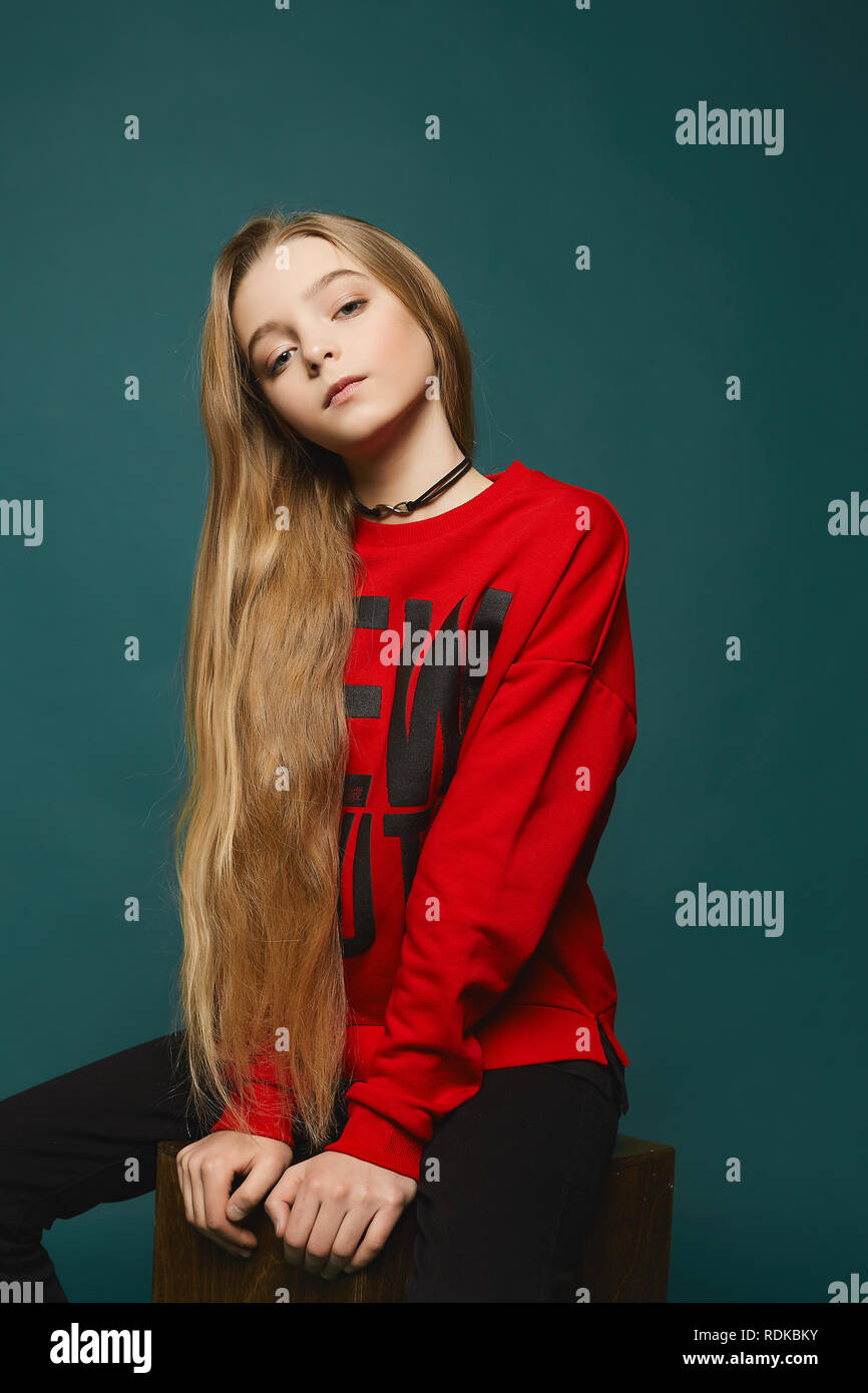 Stylish teen, beautiful young model girl with long blonde hair in black jeans and in fashionable red sweatshirt posing at the studio Stock Photo